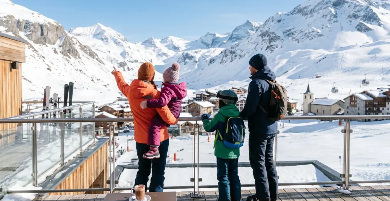 Une famille avec deux enfants en tenue de ski colorée, vue de dos, admirant le panorama montagneux enneigé depuis la terrasse d'un hôtel alpin contemporain sous un soleil d'hiver éclatant