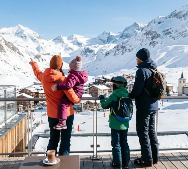Une famille avec deux enfants en tenue de ski colorée, vue de dos, admirant le panorama montagneux enneigé depuis la terrasse d'un hôtel alpin contemporain sous un soleil d'hiver éclatant